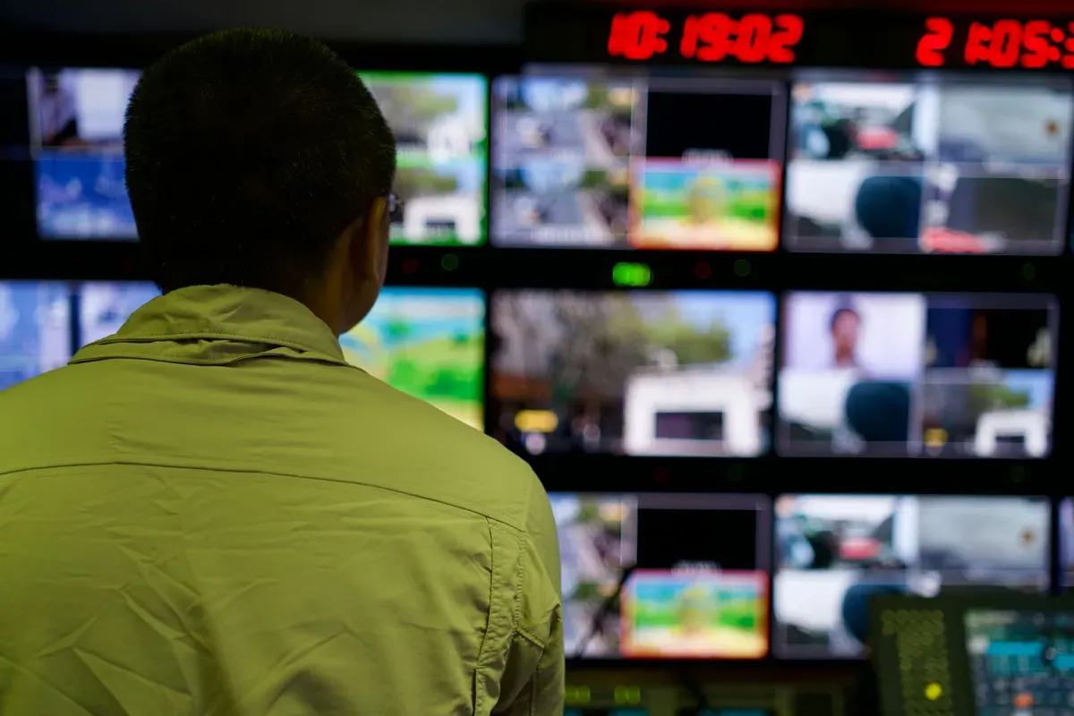 A man seen from behind watching a wall of monitors in a control room