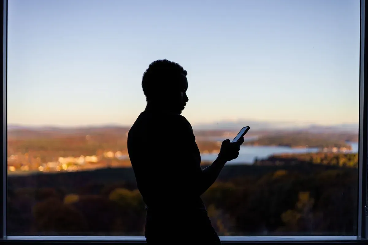 A person silhouetted against a window at dusk, looking at a phone