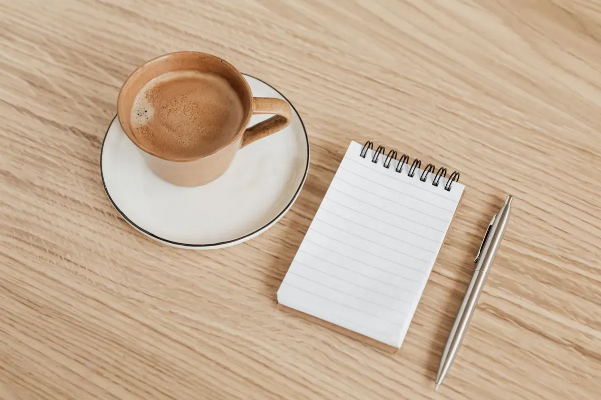 A notepad and pen beside a cup of coffee on a wooden table